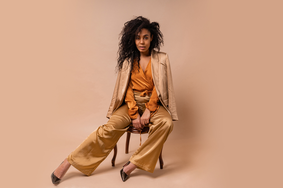 Beautiful african model with perfect curly hairs in elegant orange blouse and silk pants sitting on vintage chair onver beige background.
